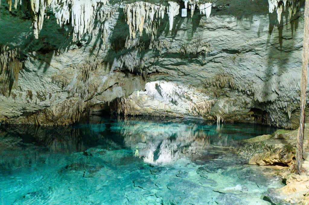 An underground cave with stalactites hanging from the ceiling and a clear blue water pool reflecting the rock formations.