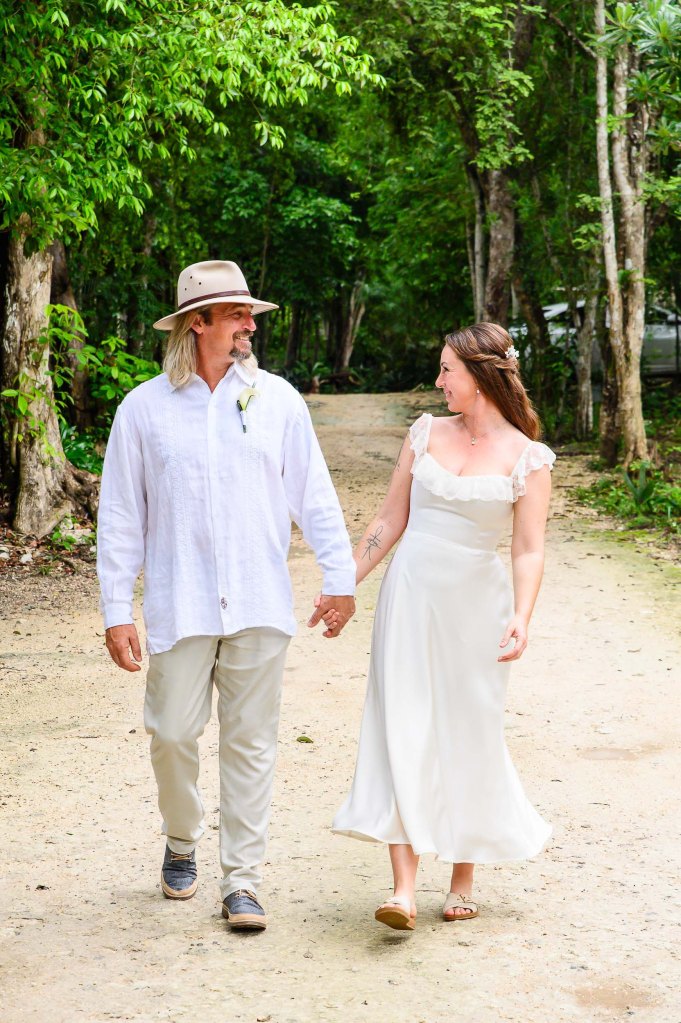 A couple walking hand in hand down a dirt path surrounded by lush greenery. The man is wearing a white shirt, beige pants, and a hat, while the woman is dressed in a flowy white gown.