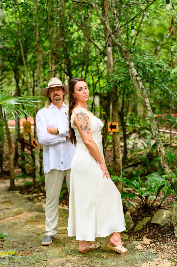 A man and woman pose together in a lush, green forest setting. The man wears a white shirt and a hat, standing with arms crossed, while the woman wears a white dress and displays a tattoo on her arm, looking back at the camera.