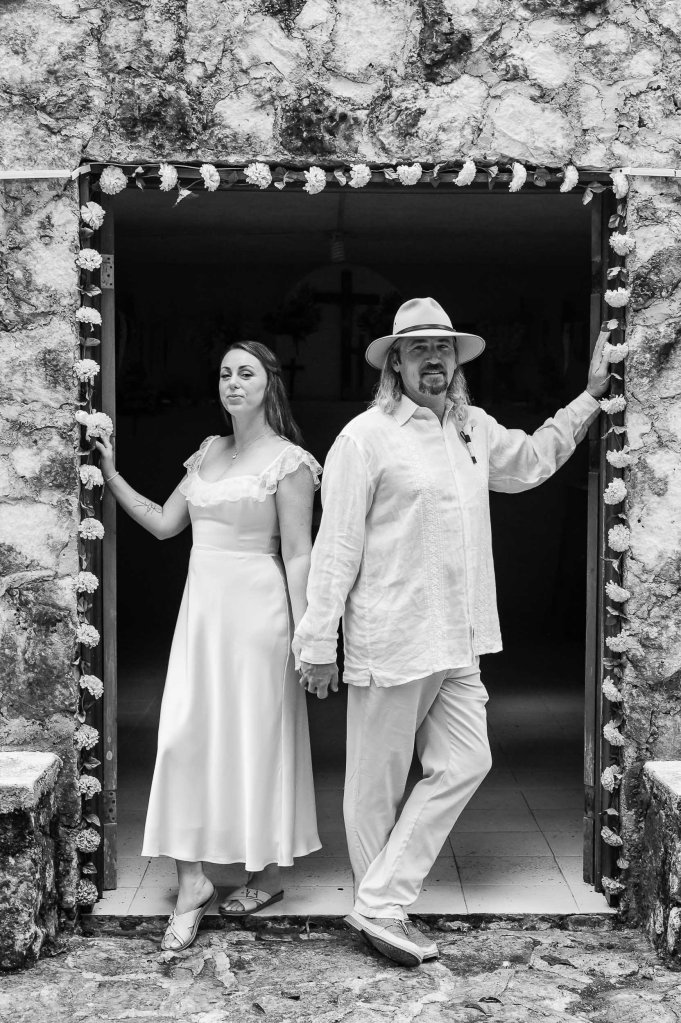 Black and white photo of a couple standing in a stone doorway, holding hands. The woman is wearing a white dress and the man is in light-colored attire and a hat. Floral decorations frame the entrance.