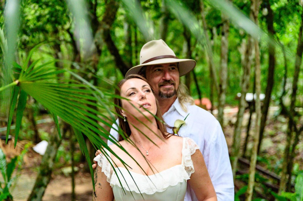A couple standing in a lush, green forest, gazing upwards with a sense of wonder. The woman wears a white dress and a necklace, while the man has a light-colored shirt and a wide-brimmed hat.