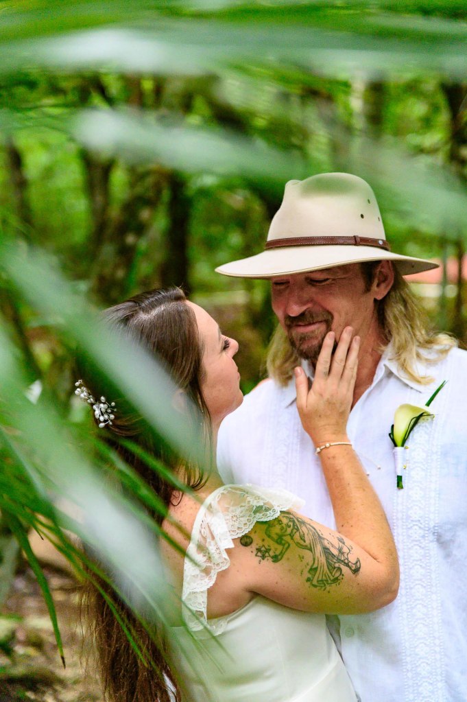 A couple embracing in a lush green forest, the woman in a white dress with intricate lace details and a tattoo visible on her arm, while the man wears a white shirt and a wide-brimmed hat, smiling at each other tenderly.