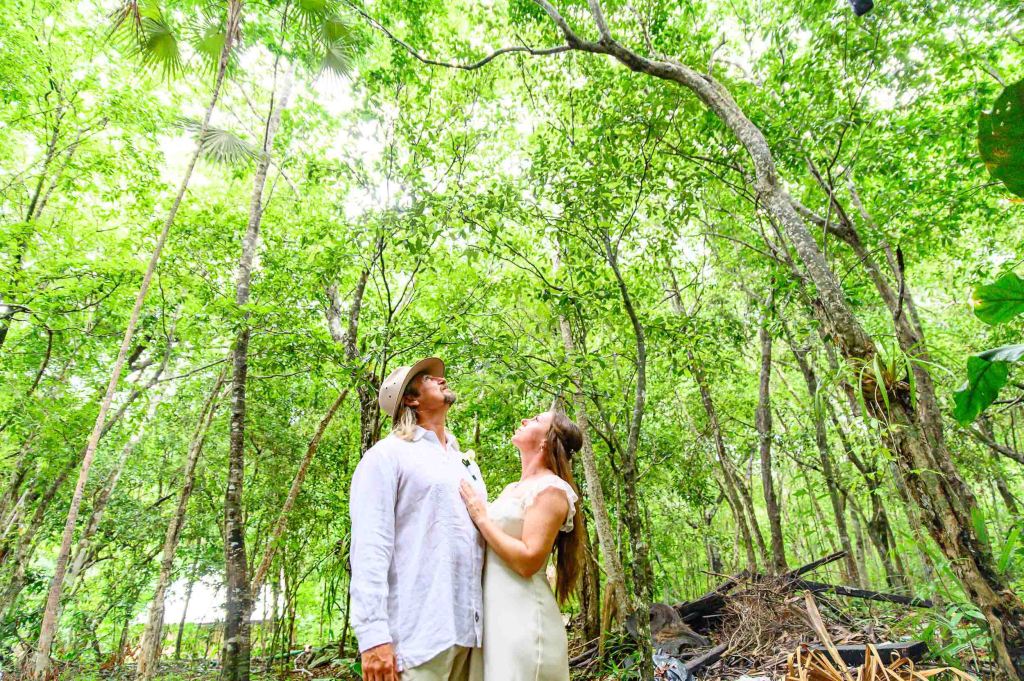 A couple standing in a lush green forest, looking up towards the trees, surrounded by vibrant foliage.
