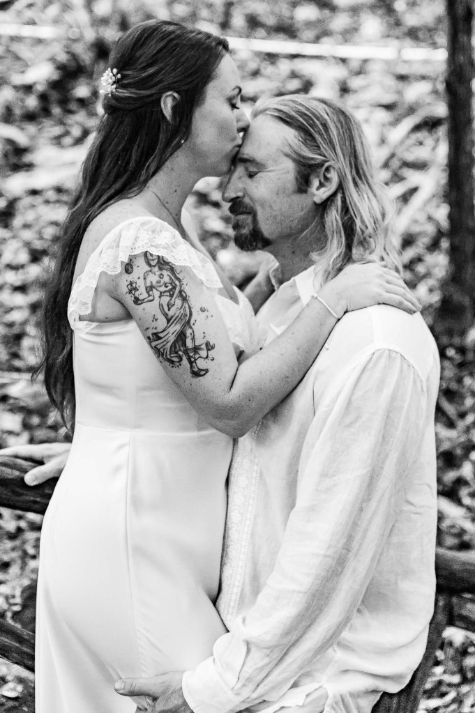 A romantic black and white portrait of a couple sharing an intimate moment in a natural setting, with the woman kissing the man's forehead while he gazes affectionately at her.