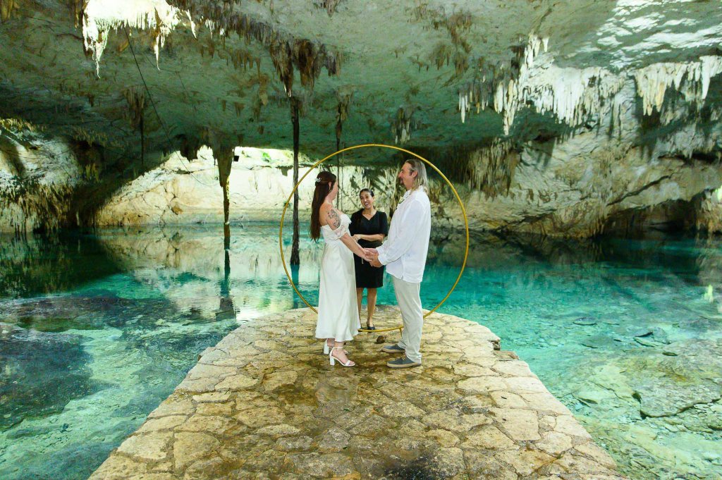 A couple standing on a stone path in a cave, holding hands during a wedding ceremony, with a circular decor behind them and water reflecting the cave's features.