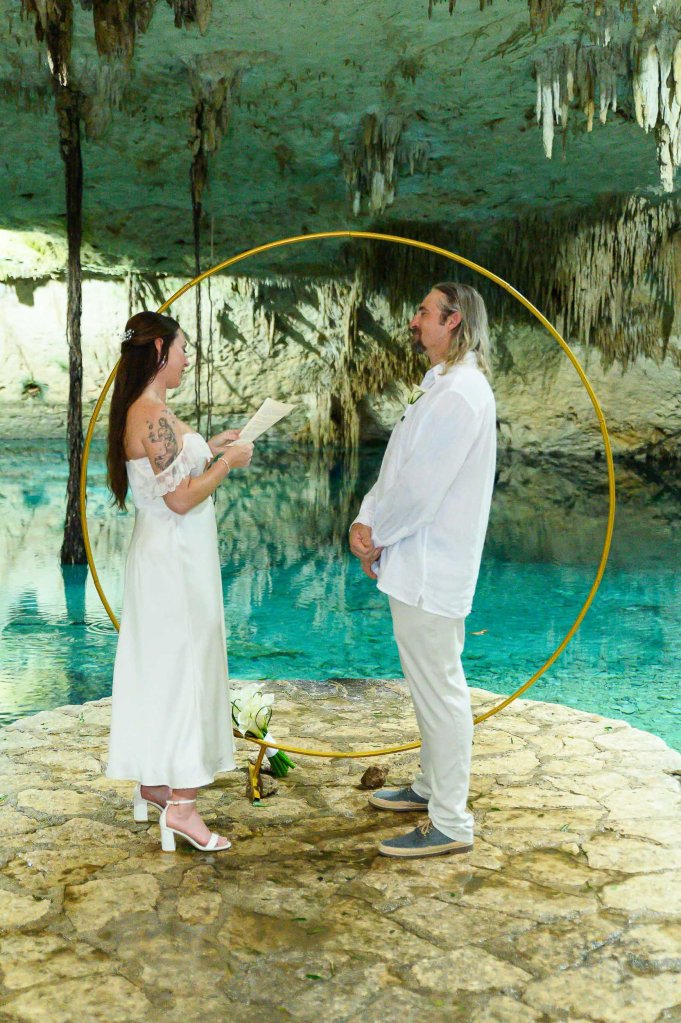 A couple stands in front of a circular floral arch in a cave setting, exchanging vows. The woman wears a white dress and the man is in a white shirt with light-colored pants. The scene features a turquoise pool and natural rock formations.