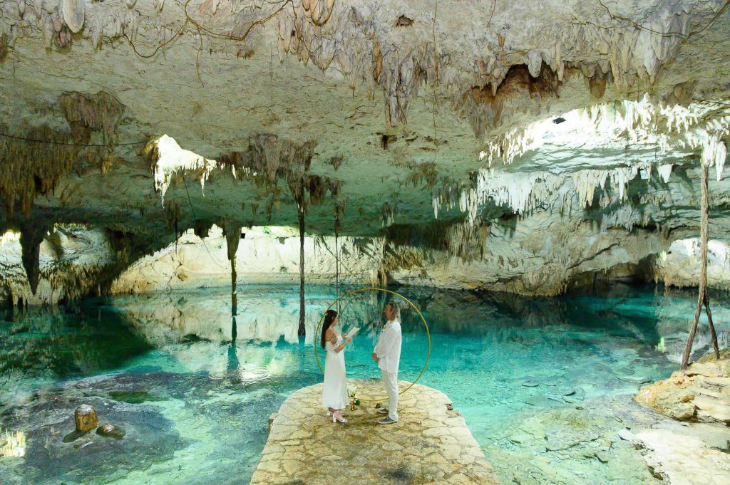 A couple standing on a stone platform in a cave, surrounded by clear blue water and towering limestone formations.