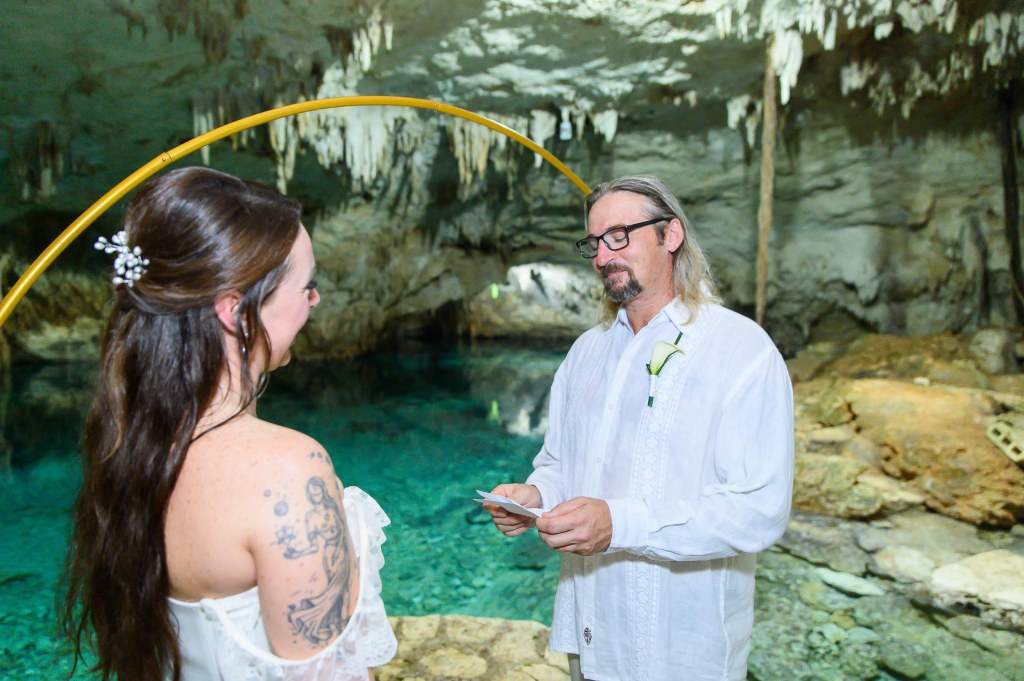 A couple standing in a cave during a wedding ceremony, with the groom reading vows and the bride listening, surrounded by natural rock formations and clear water.
