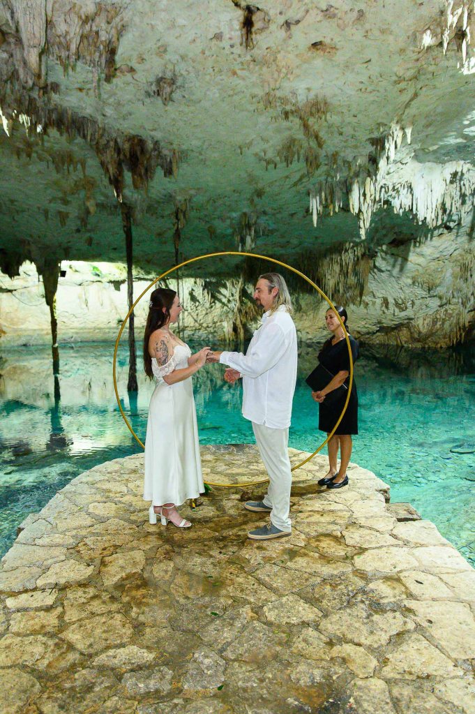 A couple exchanging vows during a wedding ceremony in a cave setting, with a circular arch in the background and a officiant present.