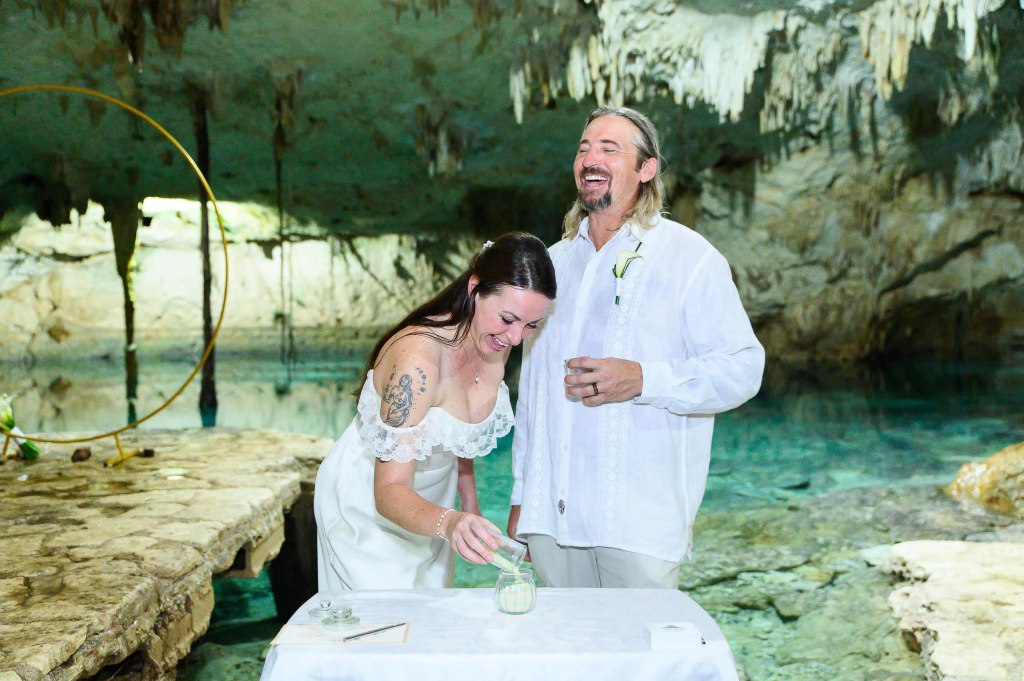 A couple laughing together during a wedding ceremony inside a cave, with a pool of water in the background and decorative elements around them.