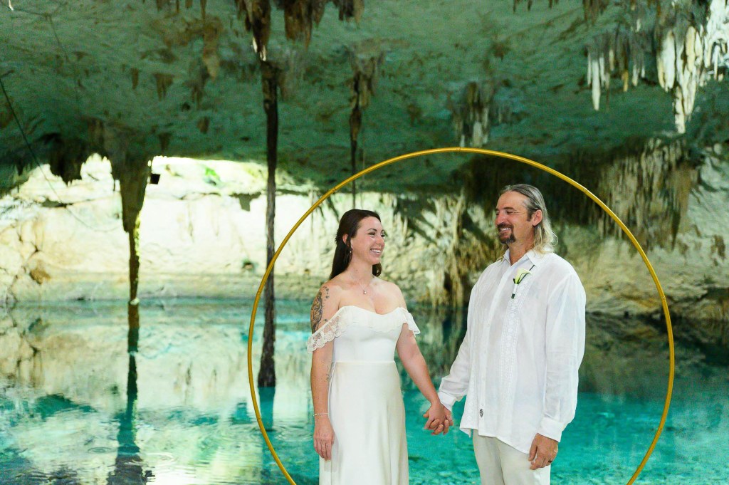 A couple holding hands in a romantic setting, standing in front of a circular frame in a cave with clear blue water and stalactites in the background.