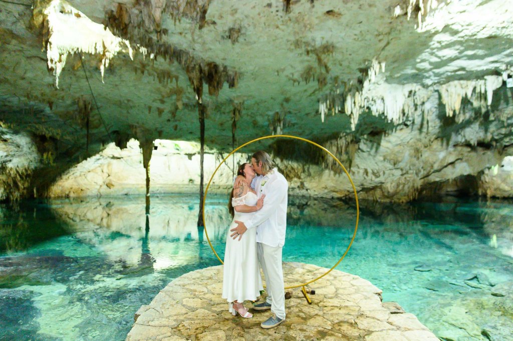 A couple embracing in a cave with stunning stalactites and clear blue water, surrounded by a natural rocky environment.