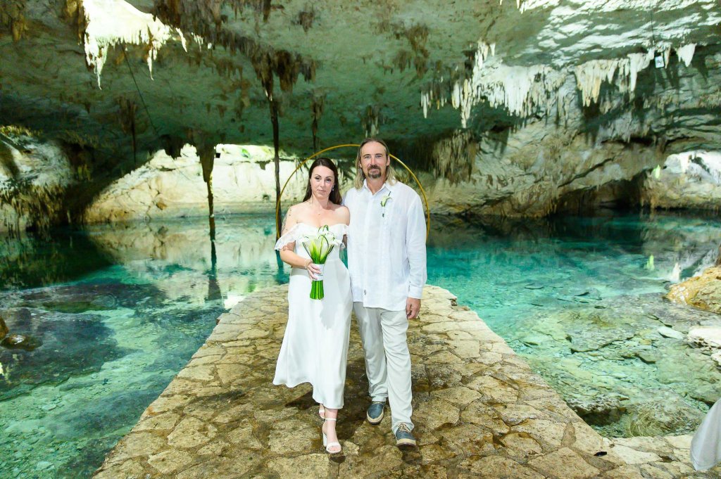 A couple walking together on a stone pathway inside a cave, near clear water, with rock formations and natural lighting.