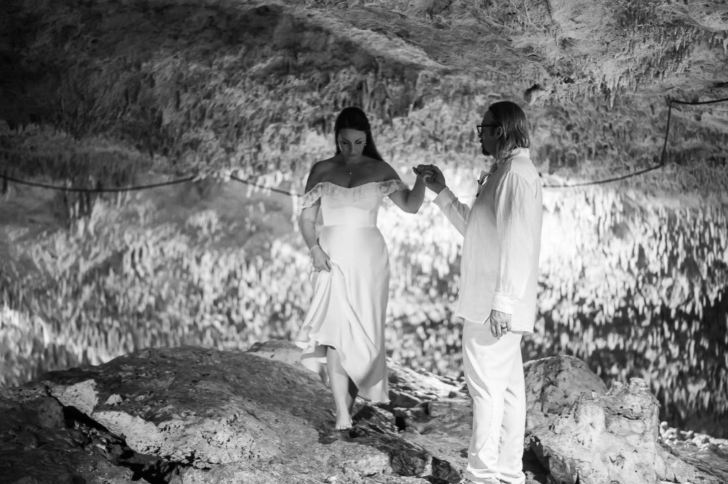 A couple dressed in elegant attire standing on rocky terrain inside a cavern, holding hands while walking together.
