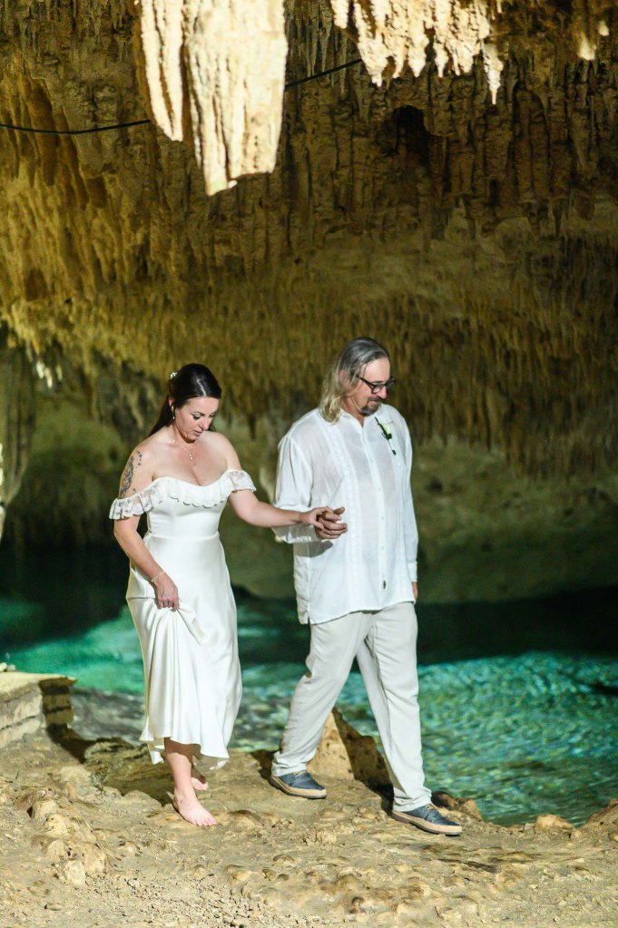 A bride in a white dress and a groom in a light outfit walk hand in hand through a cave with clear water and stunning rock formations.