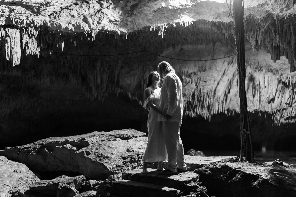 A romantic couple standing together in a cave, surrounded by stalactites and stalagmites, captured in black and white.