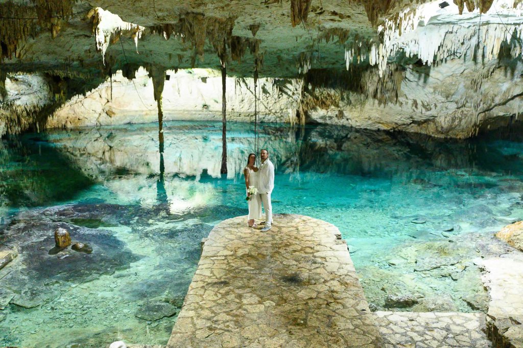 A couple stands on a stone platform in a serene underground cave, surrounded by clear turquoise water and stunning stalactites hanging from the ceiling.