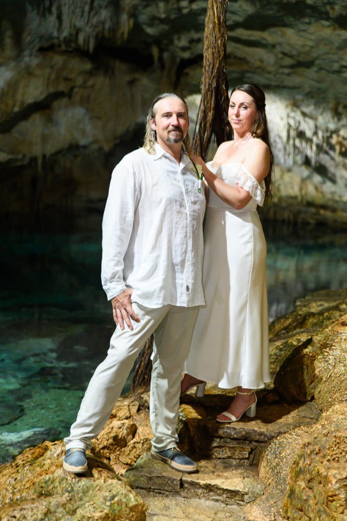 A couple poses together in a cave setting, surrounded by rocky terrain and water. The man wears a white linen shirt and light pants, while the woman is dressed in a white, off-shoulder gown.