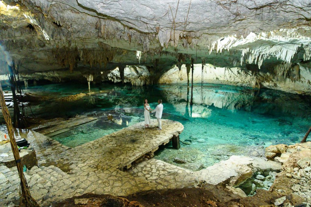 A couple stands on a stone platform in a cave, surrounded by clear blue water and stalactites hanging from the ceiling.