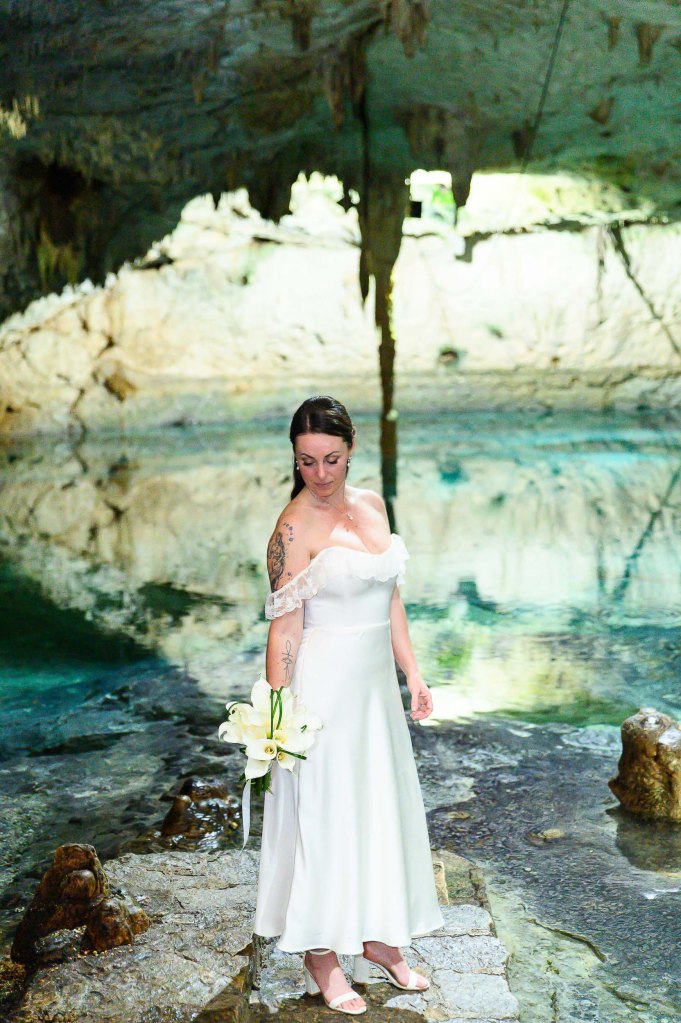 A bride in a white dress holding a bouquet of lilies stands on a stone pathway in a cave, with a serene water body and stalactites in the background.