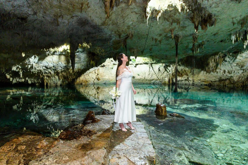 A woman in a flowing white dress stands on a rocky ledge inside a cave, surrounded by clear water and stunning rock formations.