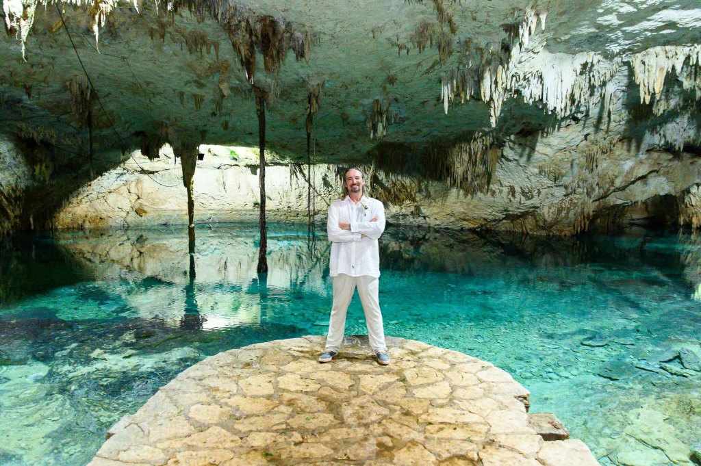 A man stands confidently on a stone platform in a cave, surrounded by clear blue water and stunning rock formations above him.