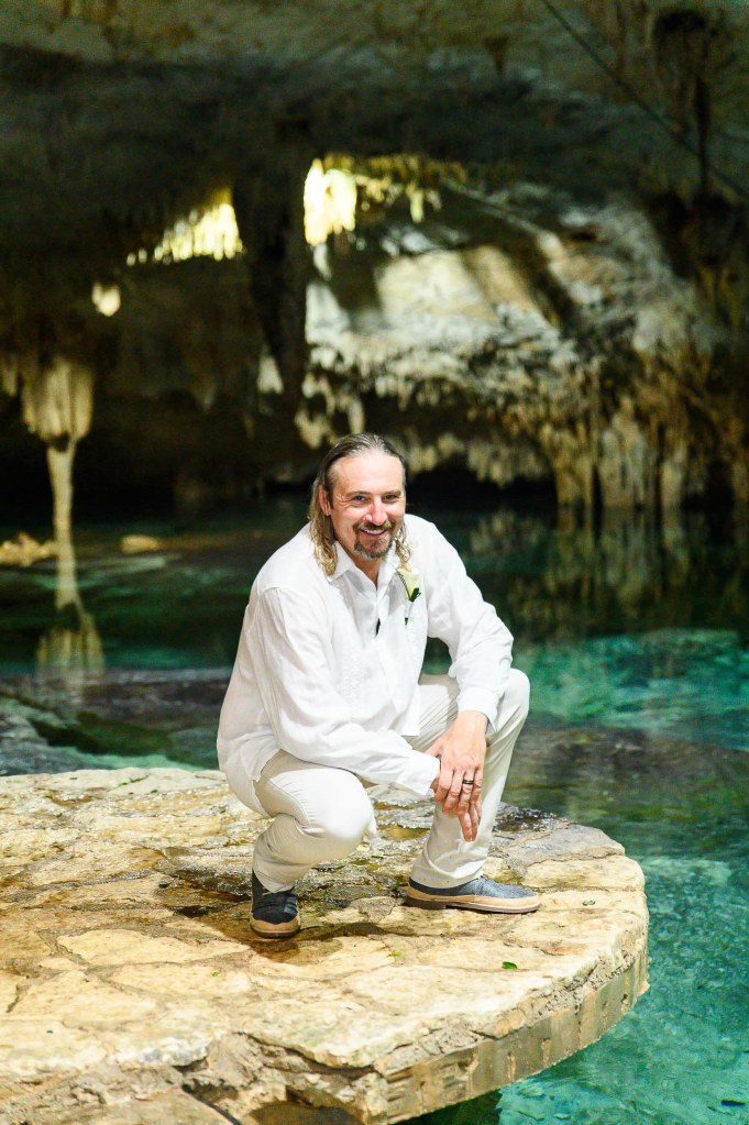 A man in a white outfit kneels on a rocky ledge above clear water in a cave, surrounded by stalactites and stone formations.